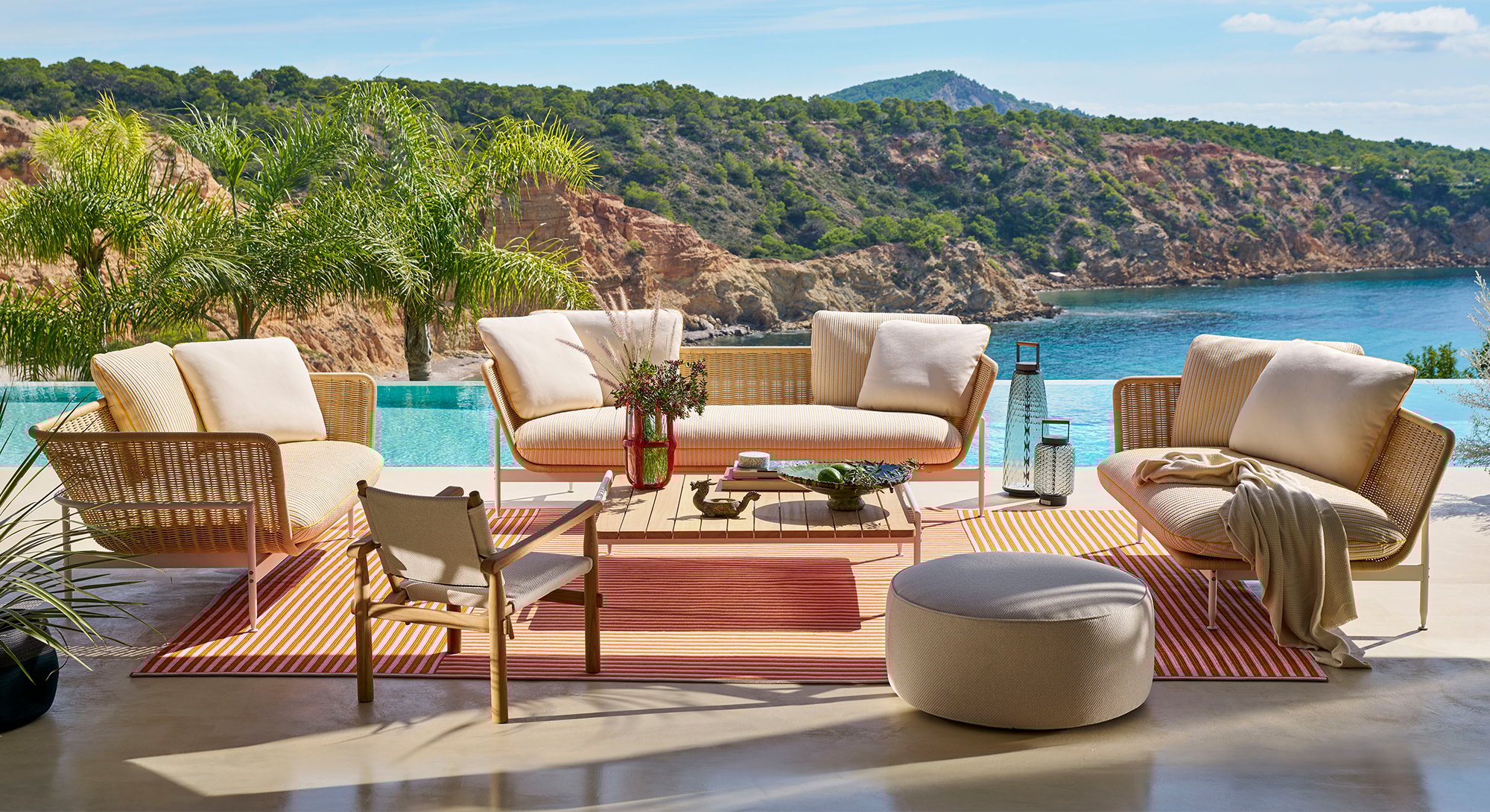 A scenic yet sustainable outdoor patio furniture with a 3 seater, a 2 seater, 1 chair and 1 pouf placed along with a wooden table overlooking lush green mountains and water.