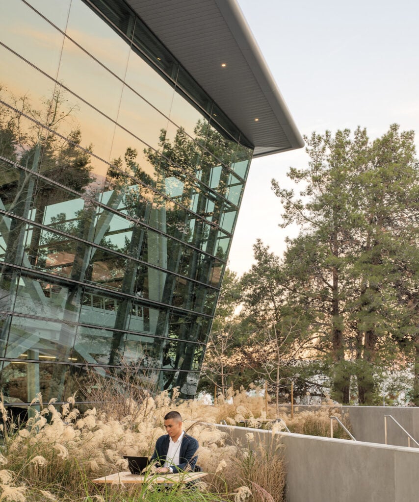 A person sitting outside for this call with a laptop embracing the native plants in the outdoor of a glass building of the NVIDIA Campus.