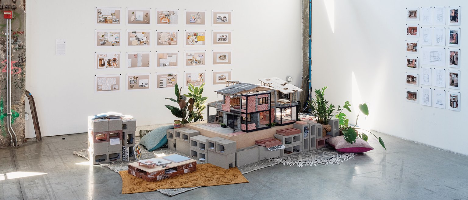 A display of the 3D model of a house at the centre of an exhibition space, placed on the floor with rugs, plants and cushion to make the space look warm.