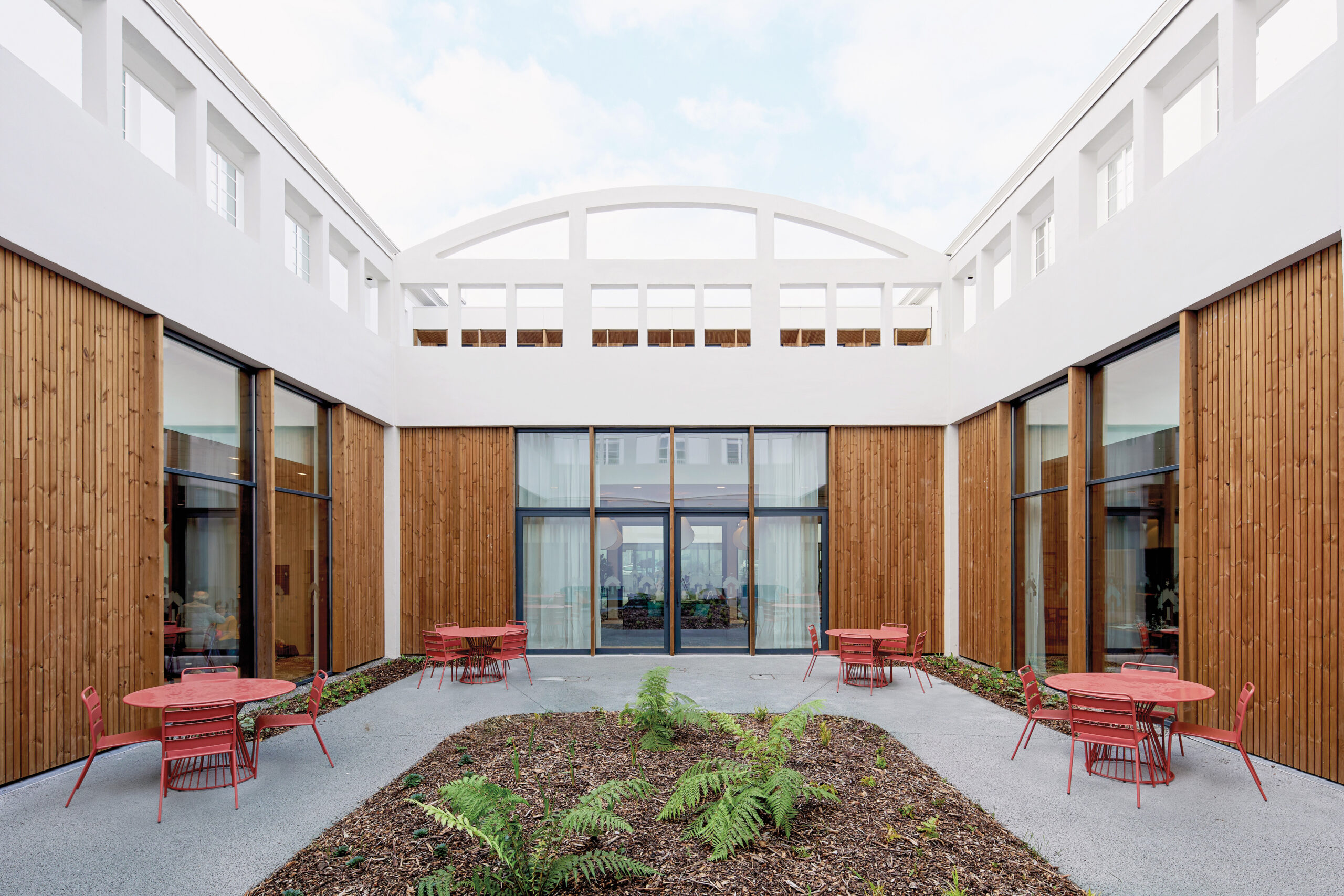 The u shaped courtyard with red outdoor furniture and wood facade walls open to sky.