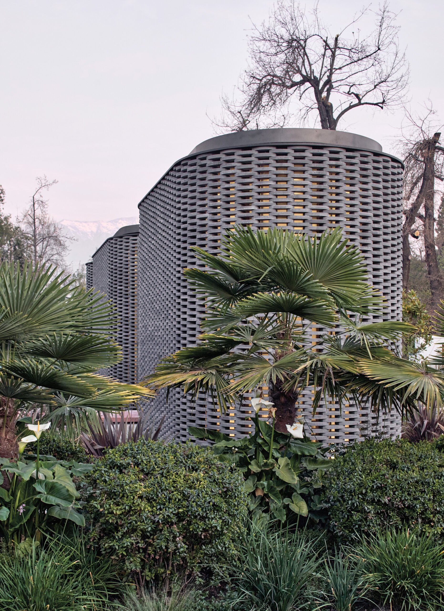 An exterior of a organic curved grey brick wall with a lot of greenery around.