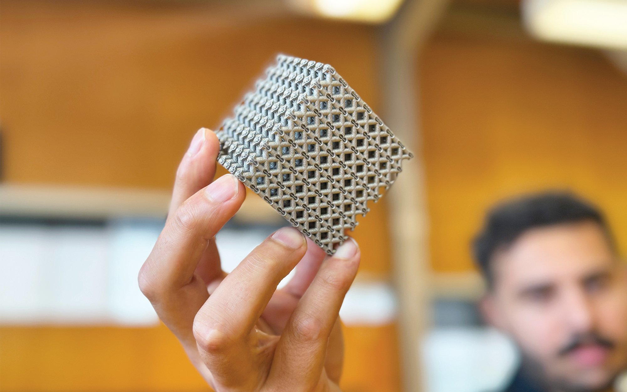 A PhD student holding a 3D-printed titanium (metal) lattice cube that is in focus.