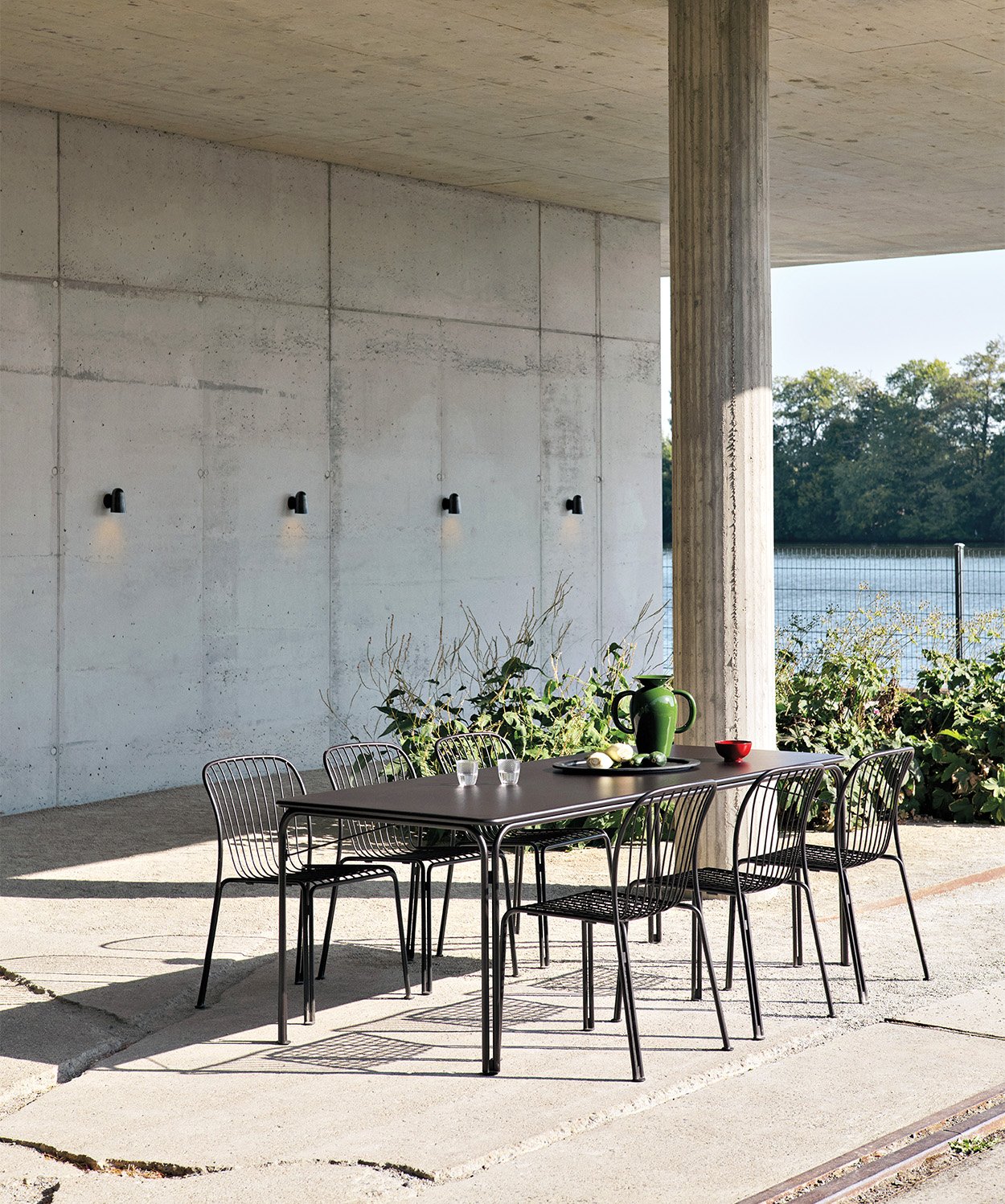 Outdoor dining set placed in t a raw concrete outdoor space with green on the edges of the space.