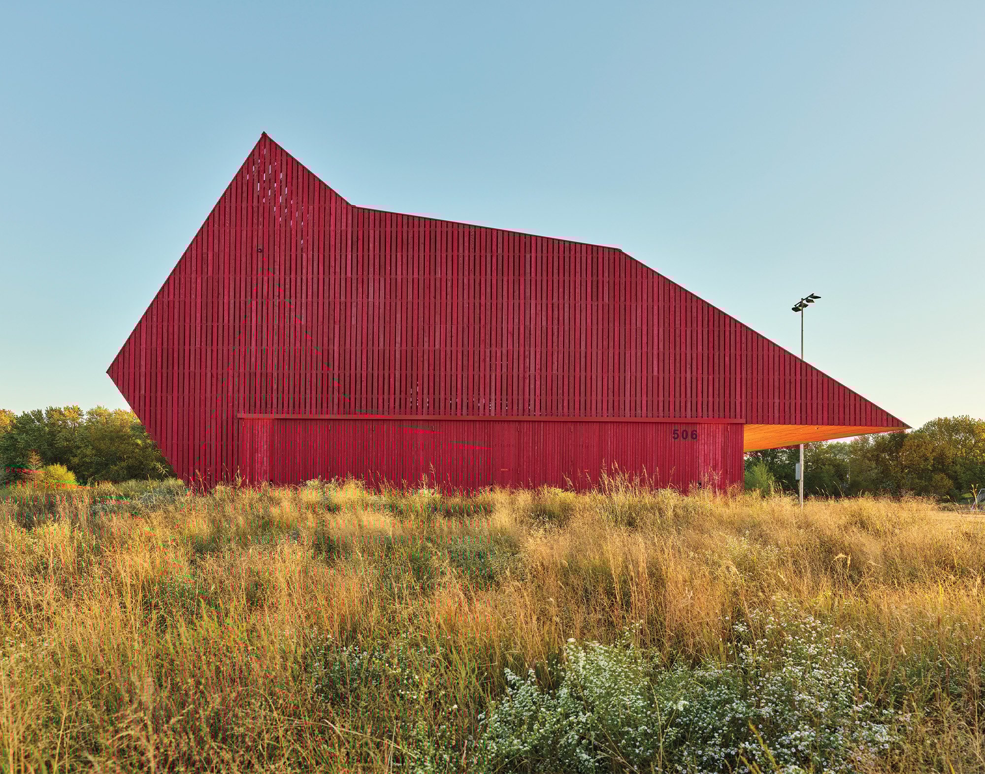A sideview of the bright red angular long building structure in the hay and grass outdoors completely made in wood.