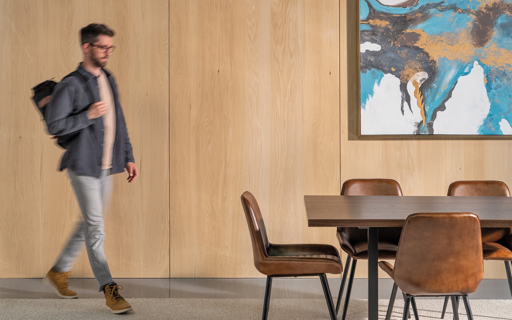 A person walking towards a dining wooden table with a backpack and the walls are covered in natural wood.